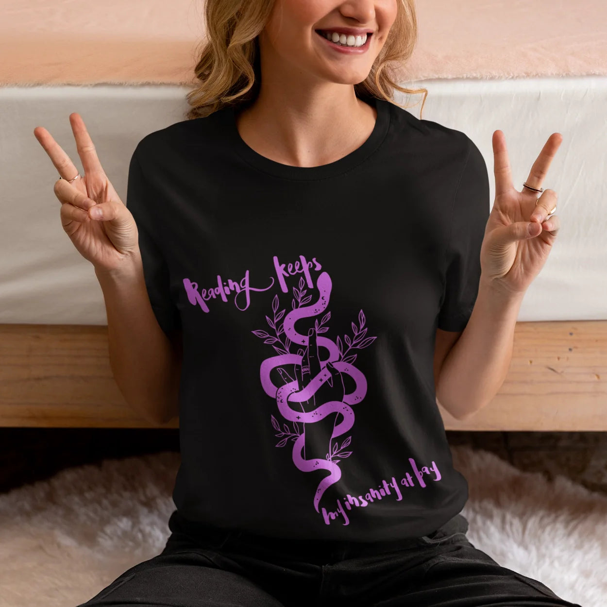 Person wearing a black t-shirt with a purple snake design and text, sitting on a bed.